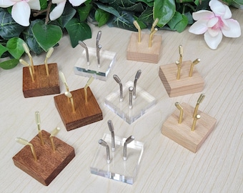 May include: A collection of square display stands featuring either clear acrylic or wooden bases in various shades. Each stand is equipped with two or three upright metal prongs with angled tips, designed to hold small objects securely in place.