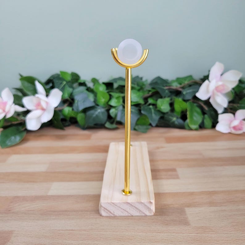 Puede incluir: Un soporte de exhibici&oacute;n con una base rectangular de madera clara y una varilla vertical de metal dorado. En la parte superior de la varilla hay una cuna semicircular dorada que sostiene un objeto esf&eacute;rico blanco, liso y opaco.
