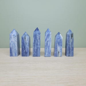 May include: Six blue kyanite crystal towers are arranged on a light wood surface. The crystals have a vertical, columnar shape with a pointed top. The blue color is streaked with white and gray.
