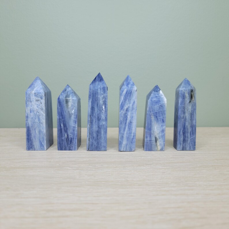 May include: Six blue kyanite crystal towers are arranged on a light wood surface. The crystals have a vertical, columnar shape with a pointed top. The blue color is streaked with white and gray.