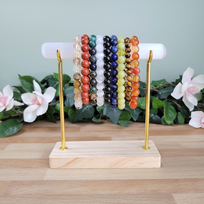 Puede incluir: Una colecci&oacute;n de diez pulseras de cuentas con diversas piedras naturales en colores que incluyen transparente, rojo, verde, blanco, negro, azul, amarillo, marr&oacute;n y naranja, exhibidas en una barra horizontal blanca con soportes dorados.