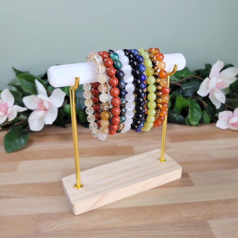 Puede incluir: Una colecci&oacute;n de diez pulseras de cuentas con piedras preciosas de varios colores, incluyendo transparente, rojo, verde, blanco, negro, azul, amarillo y naranja, exhibidas en una barra horizontal blanca con soportes de metal dorado y base de madera.