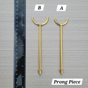 Puede incluir: Dos piezas de metal dorado etiquetadas como A y B, con una varilla recta y una parte superior en forma de U. Una regla negra a la izquierda muestra que las piezas miden aproximadamente entre 11,5 y 12,5 cm de largo desde la base hasta la punta.