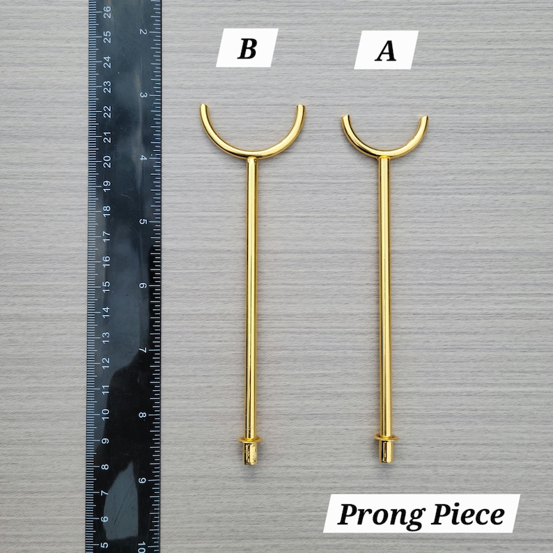 Puede incluir: Dos piezas de metal dorado etiquetadas como A y B, con una varilla recta y una parte superior en forma de U. Una regla negra a la izquierda muestra que las piezas miden aproximadamente entre 11,5 y 12,5 cm de largo desde la base hasta la punta.