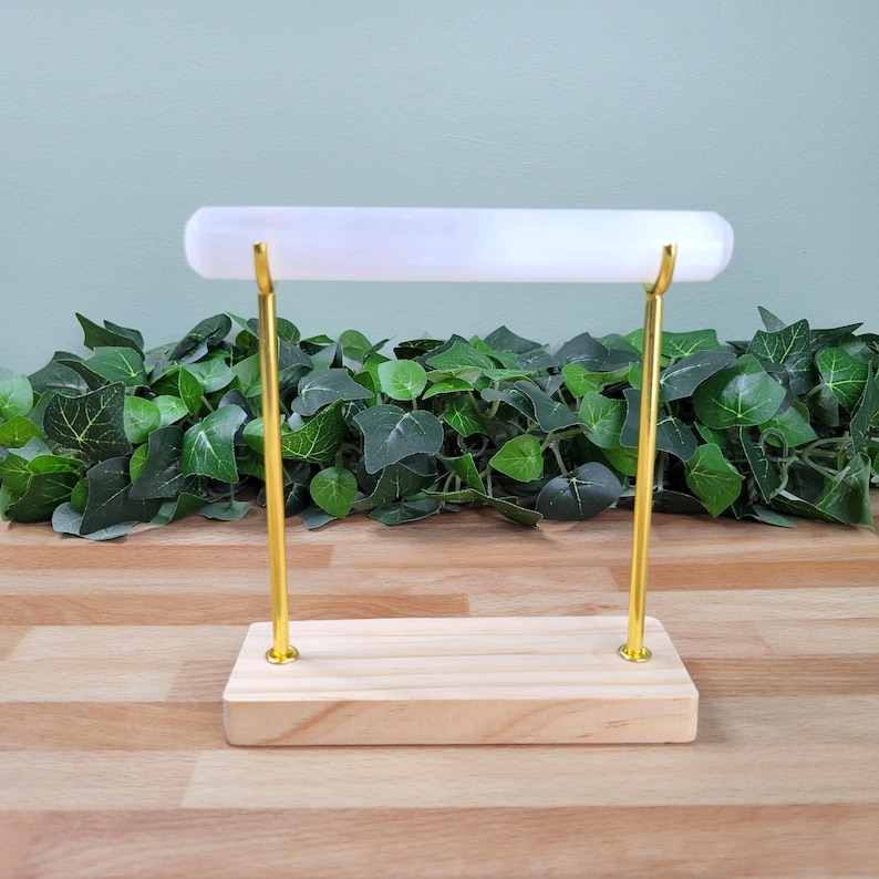 Puede incluir: Un expositor de joyas con una barra cil&iacute;ndrica blanca horizontal sostenida por dos varillas met&aacute;licas finas de tono dorado. Las varillas est&aacute;n fijadas a una base rectangular de madera clara, lo que proporciona una base estable a la estructura.