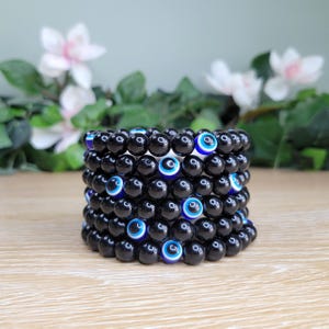 May include: Stack of black beaded bracelets with blue and white evil eye beads. The bracelets are stacked on a light brown surface with a blurred background of white flowers and green leaves.