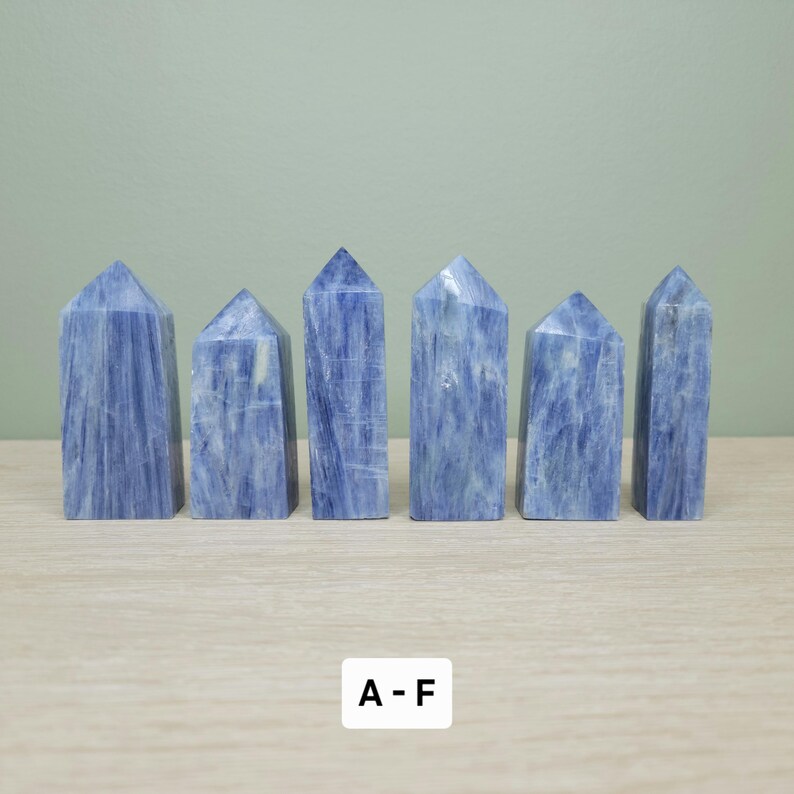 May include: Six blue kyanite crystal towers with a natural, textured appearance. Each tower has a pointed top and varying shades of blue and white. The crystals are arranged on a light-colored wooden surface.