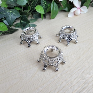 May include: Three silver-colored, crown-shaped sphere holders. Each holder has a circular opening at the top and a decorative, beaded design around the rim. The holders sit on small, rounded feet.