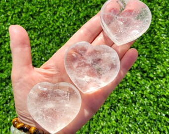May include: Three clear quartz heart-shaped stones are held in an open hand against a backdrop of green grass. The stones vary slightly in size and clarity. A bracelet with brown and green beads is visible on the wrist.