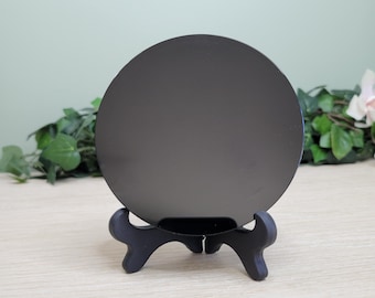 May include: A round, black object is displayed on a small, black easel. The object has a reflective surface. The easel has a three-legged design. The background features greenery and a light-colored surface.