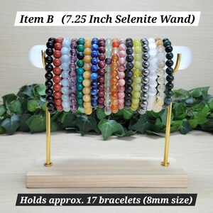Puede incluir: Un expositor con una varita de selenita de 18,4 cm que sostiene 17 pulseras de cuentas de piedras preciosas multicolores, de unos 8 mm. El soporte tiene una base de madera clara y dos brazos de metal dorado que sostienen la varita blanca.