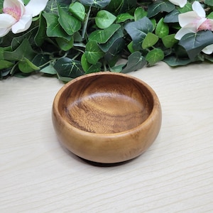 May include: A small, round wooden bowl with a natural wood grain finish. The bowl is light brown and has a smooth, polished surface. The bowl is empty and ready to be filled. The bowl is sitting on a light-colored wooden surface.
