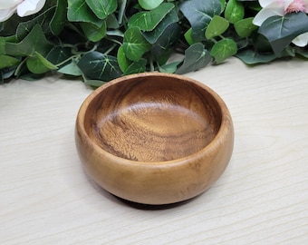 May include: A small, round wooden bowl with a natural wood grain finish. The bowl is light brown and has a smooth, polished surface. The bowl is empty and ready to be filled. The bowl is sitting on a light-colored wooden surface.