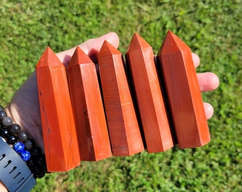 May include: Five polished red jasper crystal towers with hexagonal bases and pointed terminations. Each stone features a deep, opaque red color with subtle natural dark veining and smooth, reflective surfaces, arranged side by side in a row.