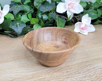 May include: A small, handcrafted wooden bowl with a natural wood grain finish. The bowl has a rounded shape with a slightly flared rim. The background includes white and pink flowers and green foliage.