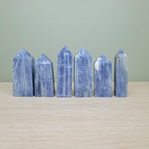 May include: Six blue kyanite crystal towers are arranged on a light-colored wooden surface against a muted green background. The crystals have a rectangular shape with a pointed top. The blue color varies in tone, with some having white streaks.
