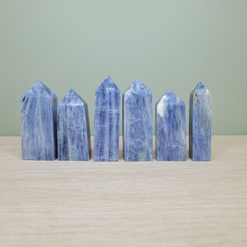 May include: Six blue kyanite crystal towers are arranged on a light-colored wooden surface against a muted green background. The crystals have a rectangular shape with a pointed top. The blue color varies in tone, with some having white streaks.