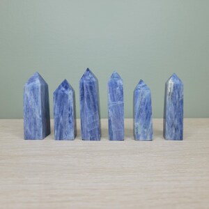 May include: Six blue crystal obelisks of varying heights are arranged on a light-colored surface. The crystals have a marbled appearance with streaks of white and blue. The background is a solid, muted green.