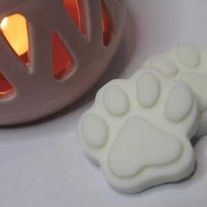 May include: Two white wax melts shaped like paw prints. The melts are on a white surface next to a pink ceramic candle warmer.