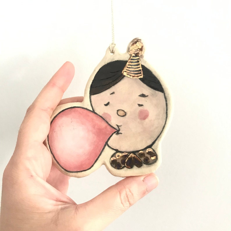 May include: A ceramic ornament of a person blowing a pink bubble with a party hat on their head. The ornament is painted with black hair, brown eyes, a pink nose, and a gold and black patterned shirt.