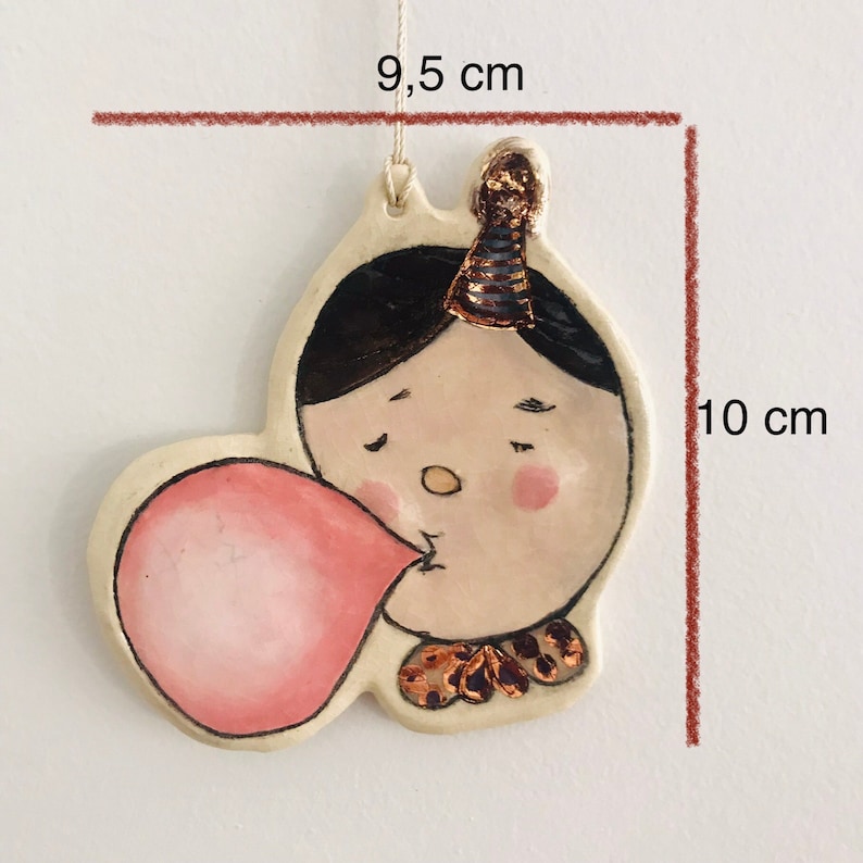 May include: A ceramic ornament of a person blowing a bubble, wearing a party hat with gold accents. The ornament is 9.5 cm tall and 10 cm wide.