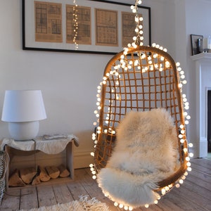 May include: A warm, inviting interior with a hanging wicker chair lit by fairy lights. The chair has a fluffy white cushion. A white lamp, a wooden crate with firewood, and a shaggy rug complete the scene. The room has wooden flooring.