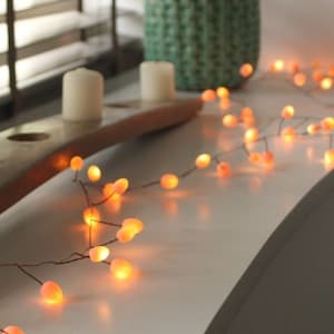 May include: A string of warm orange LED lights, resembling small glowing orbs, are draped across a white surface. The lights are part of a decorative arrangement, with a green textured vase and two white candles in the background. The scene evokes a cozy, inviting atmosphere.