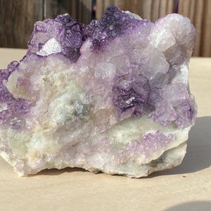 May include: A natural fluorite crystal cluster with a mix of purple and white hues. The crystal formation has a rough, textured surface with areas of translucent and opaque sections. The stone is displayed on a light-colored surface.