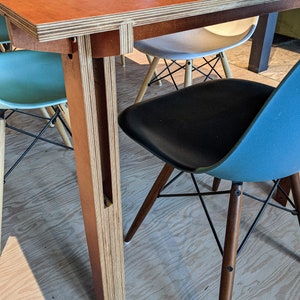 May include: A modern dining table with a reddish-brown tabletop and layered wood legs. The table is paired with several colorful chairs, including a black seat with wooden legs and a blue seat.