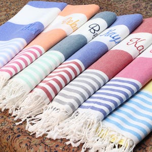 May include: A set of six striped beach towels with fringe edges. Each towel has a different color and is personalized with a name: Alex, Olivia, Bailey, Kay, Kara, and Melissa.