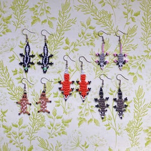 Può includere: Una collezione di orecchini a forma di lucertola in perline di vari colori, tra cui nero, bianco, rosso, rosa e marrone. Gli orecchini sono esposti su uno sfondo verde chiaro con un motivo a foglie.