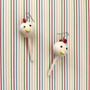 May include: A pair of white chicken-shaped earrings with red accents. The earrings are on a striped background.