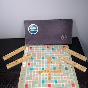 May include: Vintage Scrabble board game with wooden letter tiles. The game board is open, displaying a grid with letter and word score spaces. The box is dark purple with a circular logo. Letter holders spell out "RETRO YA", "FAMILY", "WORDS XO", and "SPELL IT".