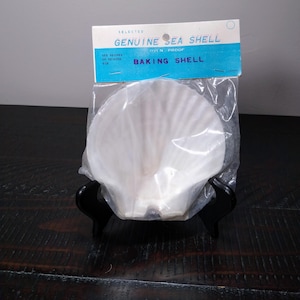 May include: A white, fan-shaped baking shell is packaged in clear plastic. The packaging includes a blue and white label that reads "GENUINE SEA SHELL BAKING SHELL". The shell is displayed on a black stand against a grey background.