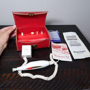 May include: A vintage red leather case with a white electric manicure set. The set includes a handheld tool with a cord, a small white box, and a set of attachments. The case has a gold latch and the tool has the word "Dominion" printed on it. The case is open and the attachments are visible. The set also includes instructions for use.