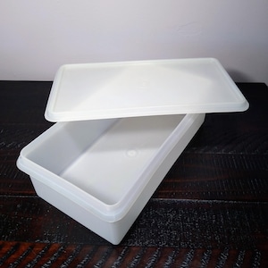 May include: A white rectangular plastic storage container with a matching lid. The lid is slightly ajar, revealing the interior of the container. The container is empty and sits on a dark wooden surface. The container is made of a translucent plastic.