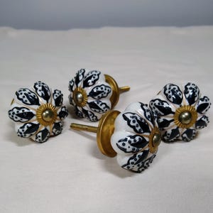 May include: Four round cabinet knobs featuring a white ceramic base with a black leaf-like pattern. Each knob has a central brass-colored metal cap and a threaded brass-colored metal bolt extending from the back for installation purposes.