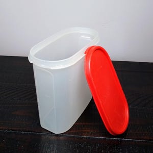 May include: A clear, rectangular plastic container with a red lid. The container is open, revealing the interior. The lid is oval-shaped and rests beside the container. The container is likely for food storage.