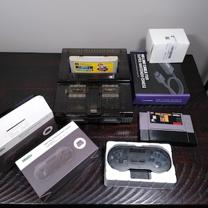 May include: A collection of retro gaming items, including a Super Nintendo console, game cartridges, a wireless controller, and an HD Link Cable. The console is black, and the game cartridges have colorful labels. The controller is black and gray.