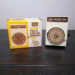 May include: A vintage Sears 24-hour plug-in light and appliance timer. The timer is white with a brown dial and black knob. The box is yellow and white with the text "Sears 24 Hour Plug-in Light and Appliance Timer."