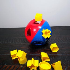 May include: A red and blue spherical shape sorter toy featuring various cutouts. A yellow cube-shaped block is placed on top, while several other yellow geometric blocks, including a star, a cylinder, and a triangle, are arranged in front.