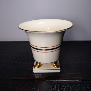 May include: Cream-colored vase with a flared top and gold trim. The vase features two horizontal bands of red and gold stripes around the middle. The base is square with gold-colored feet.