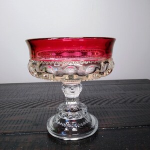 May include: A clear glass pedestal bowl with a ruby red rim. The bowl has a decorative, textured pattern around the base and a detailed stem. The bowl is sitting on a dark wooden surface.