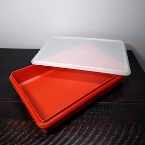 May include: A red plastic rectangular container with a white lid. The container is open, revealing the interior. The lid is slightly ajar, resting on the edge of the container. The container is likely used for food storage.