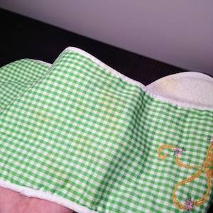 May include: Green and white gingham patterned fabric with a white border. The fabric has a yellow stain. Embroidered on the fabric is a gold-colored design with pink and blue flowers. The fabric is folded.