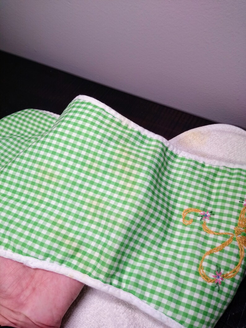 May include: Green and white gingham patterned fabric with a white border. The fabric has a yellow stain. Embroidered on the fabric is a gold-colored design with pink and blue flowers. The fabric is folded.