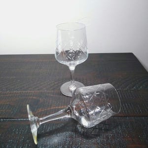 May include: Two clear crystal wine glasses with diamond-patterned etching. One glass stands upright, while the other rests on its side. The glasses are on a dark wooden surface, with a white background.
