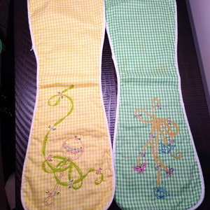 May include: Two oven mitts with a yellow and green gingham pattern. The yellow mitt features an embroidered rattle design with floral accents. The green mitt has an embroidered bow design with butterflies and birds. Both mitts have a white trim.