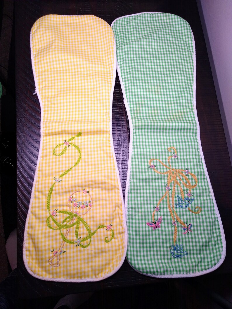 May include: Two oven mitts with a yellow and green gingham pattern. The yellow mitt features an embroidered rattle design with floral accents. The green mitt has an embroidered bow design with butterflies and birds. Both mitts have a white trim.
