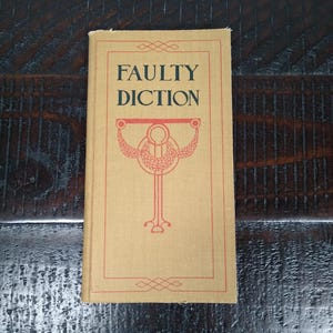 May include: A vintage book titled "FAULTY DICTION" with a beige cover and red decorative border and illustration. The book is set against a dark wooden surface.